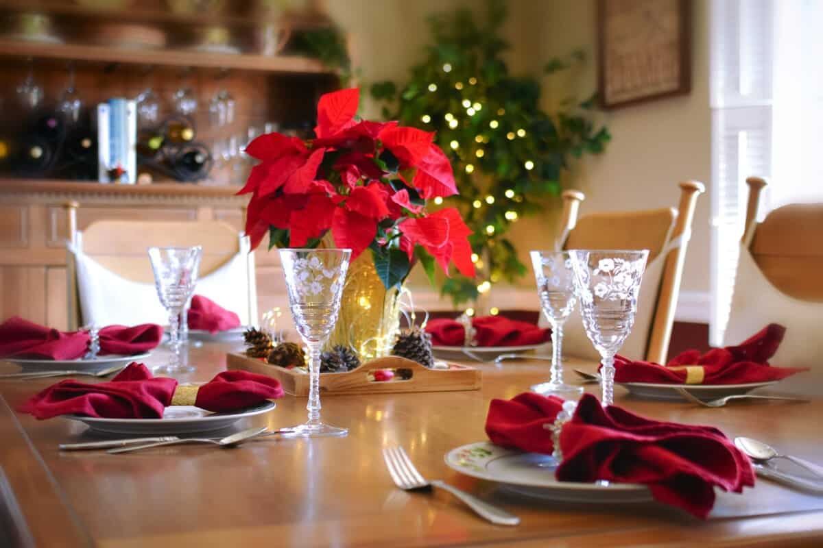 Wooden table with poinsettia flowers as centerpeice. White dishes with red napkins Pinecones surrounding Poinsettia. Fairy lights at base of Poinsettia and also on house plant in background. festive champagne glasses