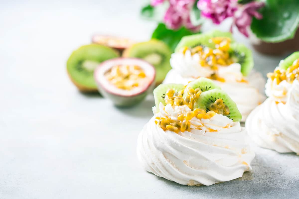 Two mini white pavlova topped with whipped cream, slices of kiwi fruit and passion fruit pulp and seeds.