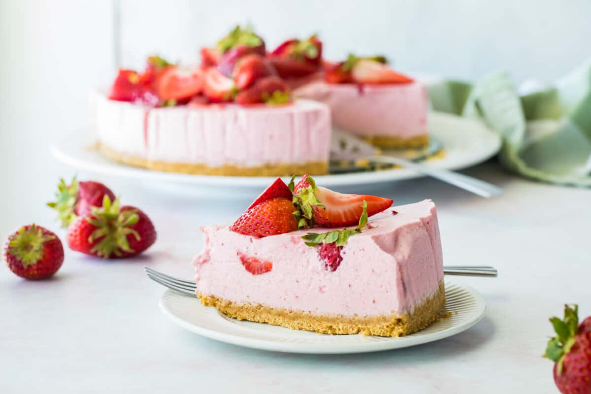 Slice of pink colored strawberry cheesecake with fresh strawberries on top and a fork on the side. The whole strawberry cheesecake is in the background on a larger plate.
