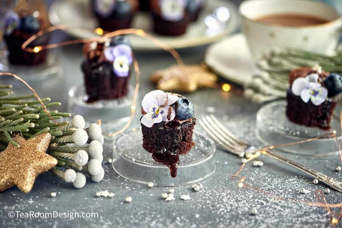 These chocolate and ginger mini cakes, a Christmas tea party menu item, are placed on clear small stands, decorated with blueberries and edible flowers