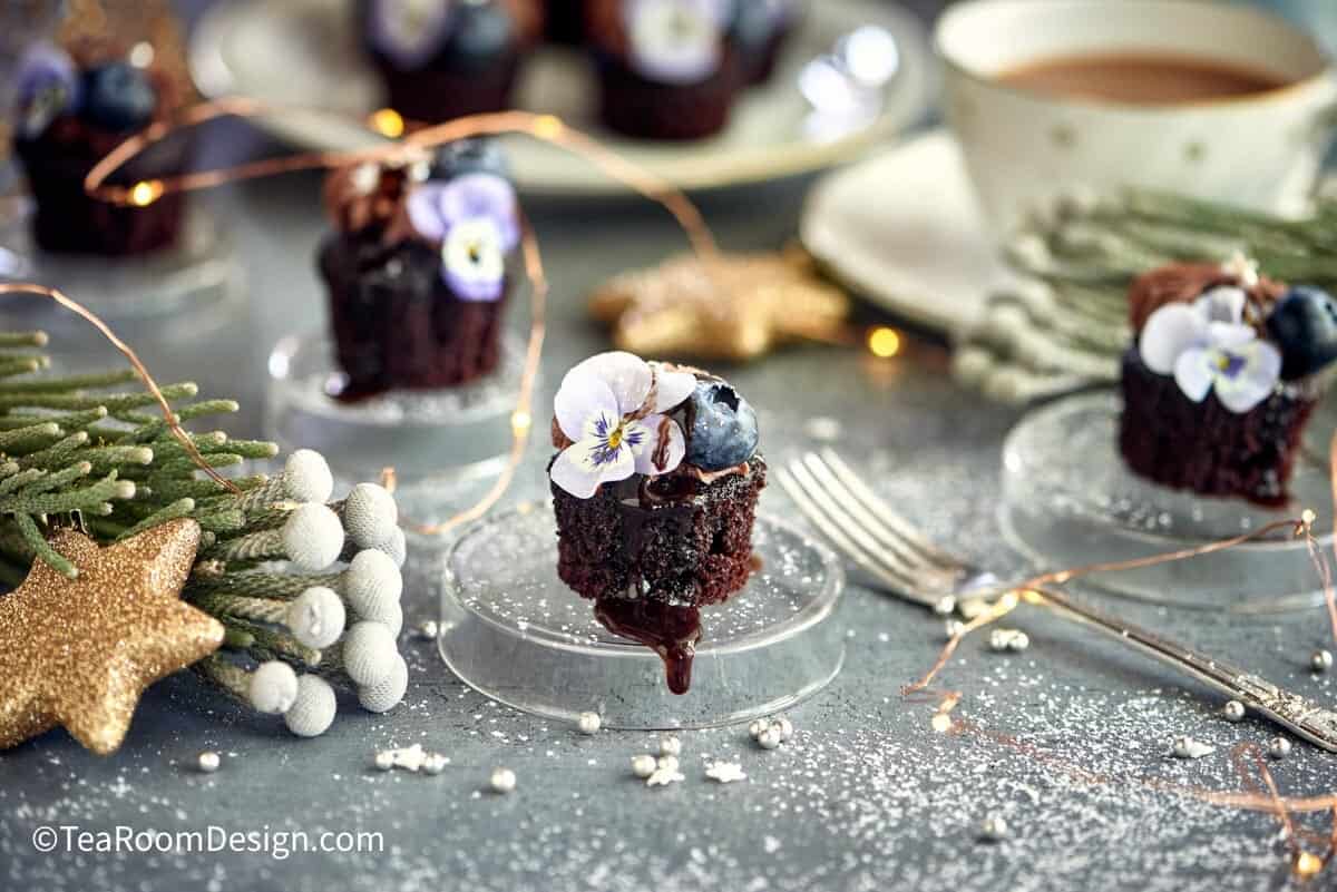 Mini chocolate cake topped with edible violet and blueberry, placed on small clear stand