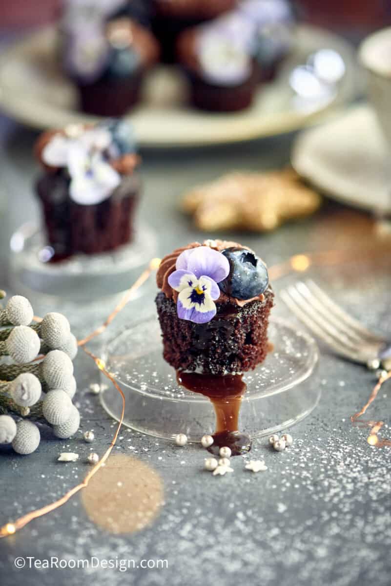 Mini chocolate cake topped with chocolate buttercream, edible violet and a blueberry, placed on a clear plastic small round platter.