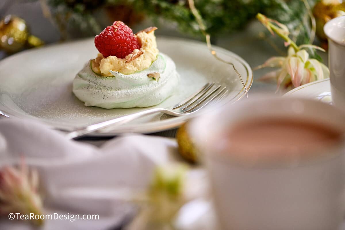 Mini light green pavlova topped with whipped cream and single raspberry on white plate with fork, placed on table with cup of tea in the foregorund.