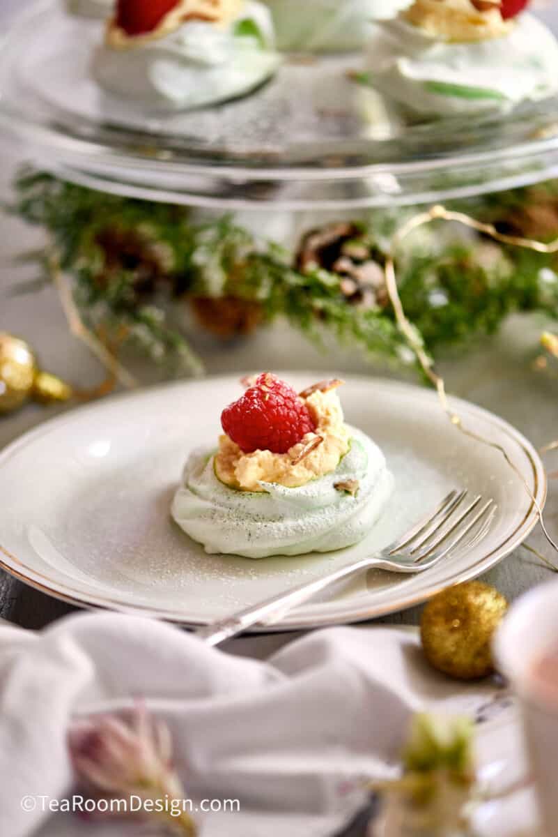 Mini green pistachio meringue topped with clotted cream and fresh raspberry on a white plate with a silver fork. Similar meringues on a clear cake stand in the background.