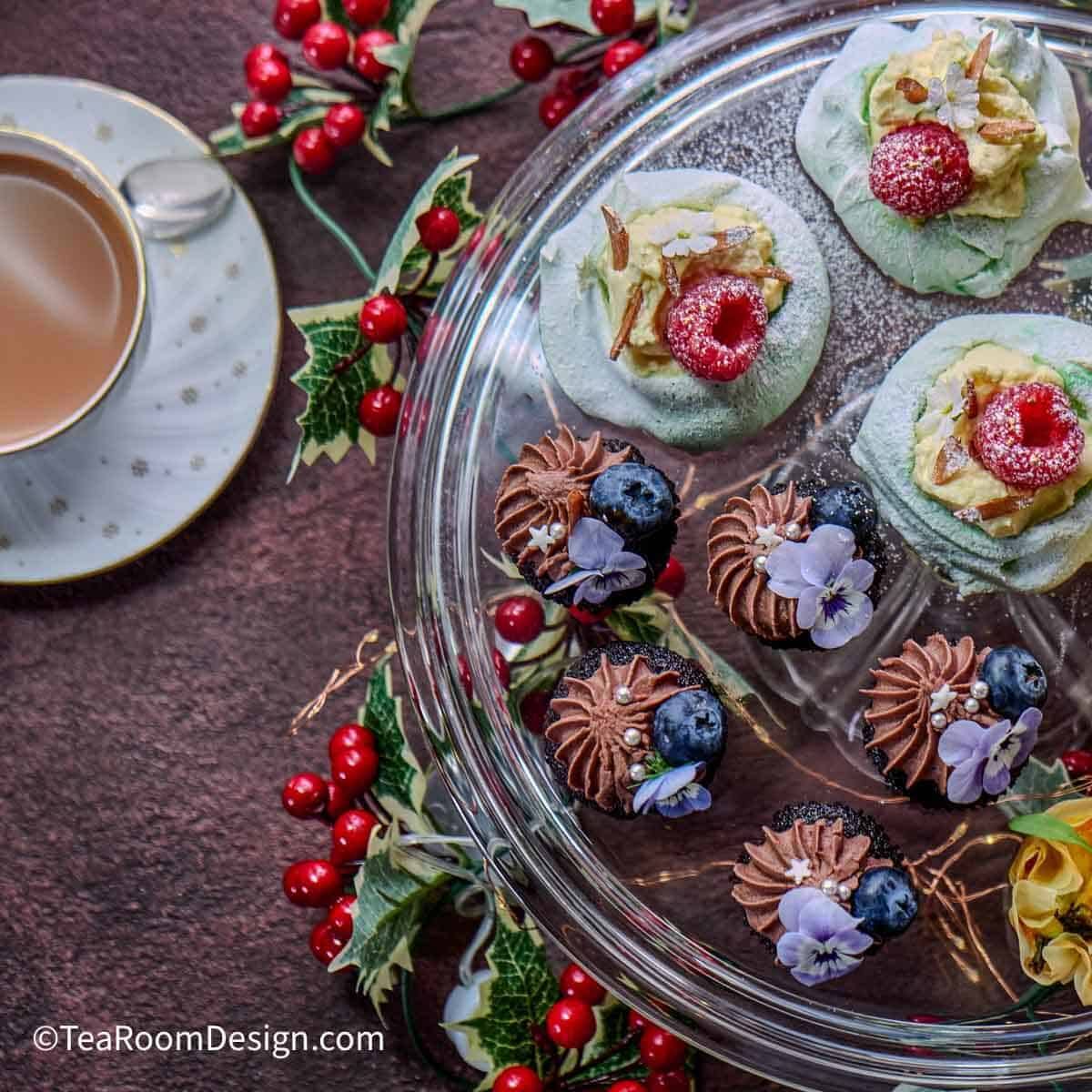 Cup of tea on left and clear round tray with examples of Christmas tea party menu items such as mini light green pavlovas topped with a fresh raspberry, mini chocolate muffins topped with a violet. Red and green table decorations.