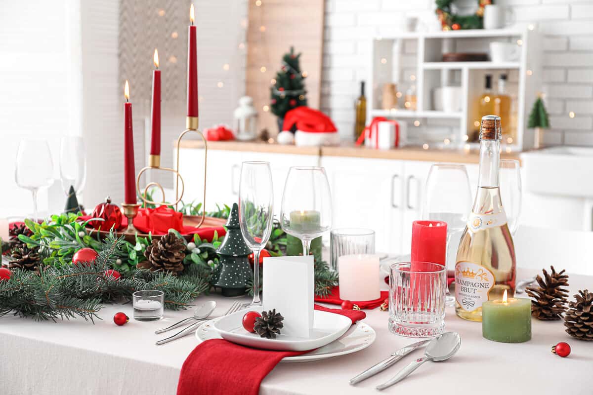 Christmas table setting. White tablecloth, white dishes, cardinal red napkins, clear glasses and champagne glasses, gold thin candlestick holder holding three red candles, surrounded by wreath made up of pine needles and pine cones. Bottle of champagne on the table and another red and green lit candle. Blurry kitchen in the background.