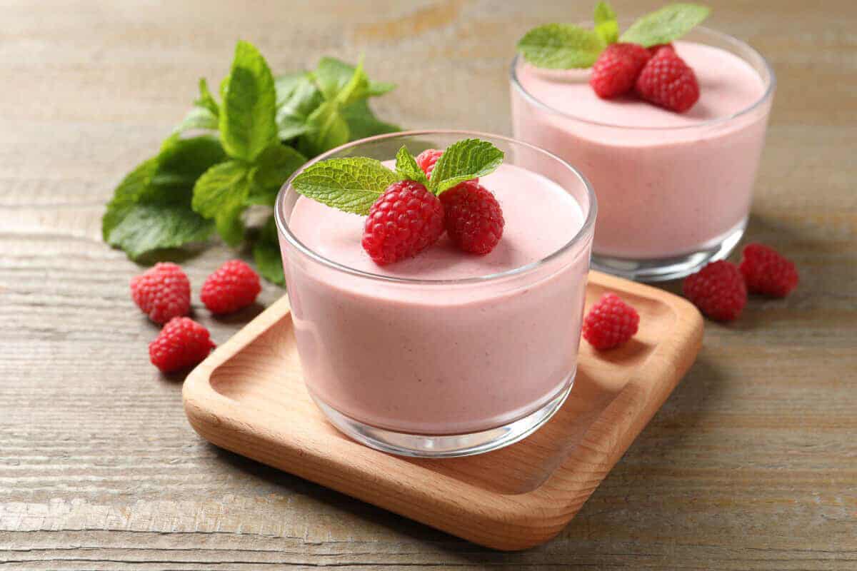 Two small clear glasses filled with pink raspberry mousse and topped with three fresh raspberries and mint leaves. One glass is on a small wodden platter.