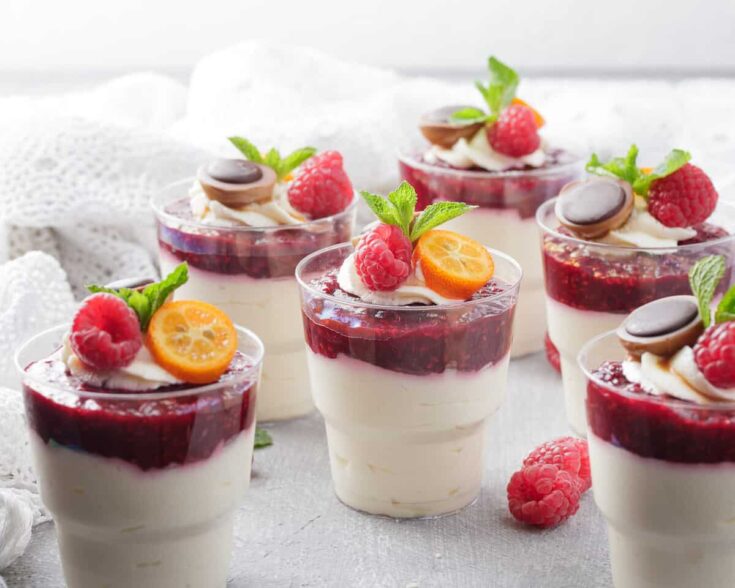 Five clear glasses filled with white panna cotta topped with layer of raspberry coulis and also some decorative raspberries and orange fruit.