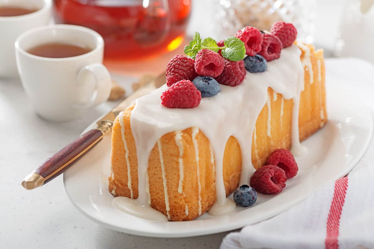 Butter vanilla loaf cake with drizzled frosting and fresh raspberries and blueberries on a plate with a clear teapot in background