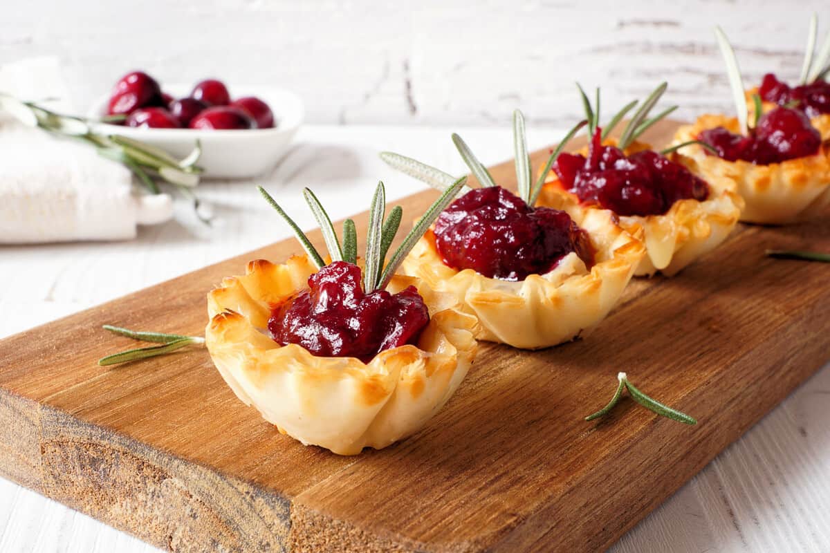 Cranberry, brie and rosemary in mini phyllo dough bites on wooden platter with cranberries in background. Lovely Christmassy colours that go well with the Christmas tea party menu.