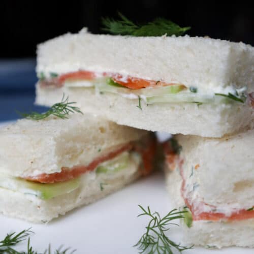 Three smoked salmon, cream cheese and cucumber tea sandwiches on a white plate with dill garnish.