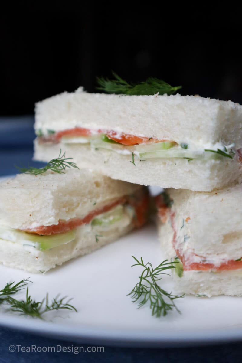 Three smoked salmon, cream cheese and cucumber tea sandwiches on a white plate with dill garnish.