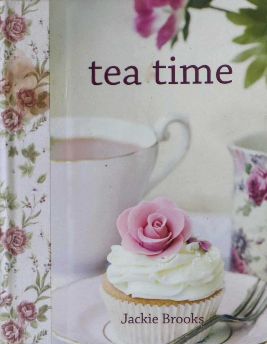 Cover of book titled "tea time" by Jackie Brooks showing cupcake with cream frosting and sugar rose on top. There is a cup of tea in the background.