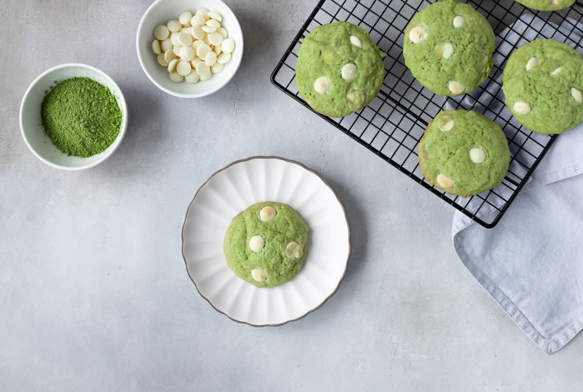 Matcha and white chocllate chip cookie on small white plate and four other cookies on cooling rack. White chocolate chips and matcha powder in two small bowls.