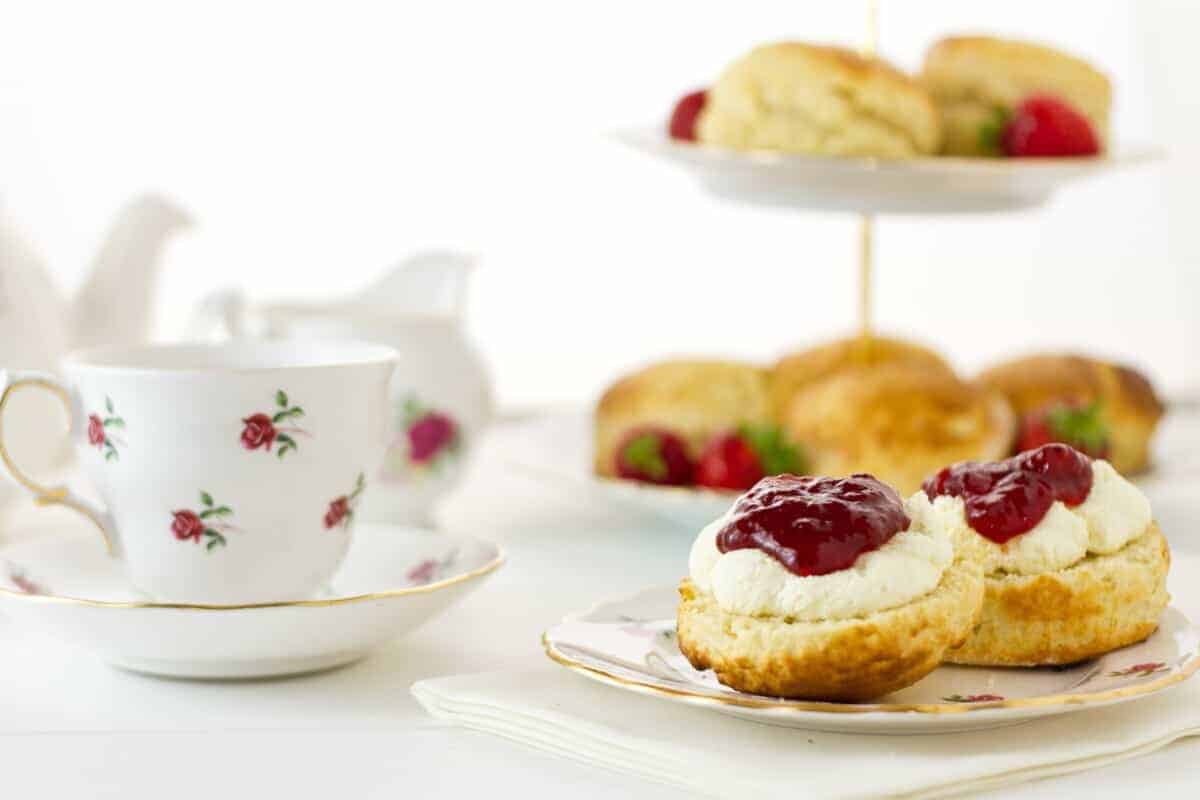 Scone with cream and strawberry jam on plate with tea cup and tea party stand in the background.